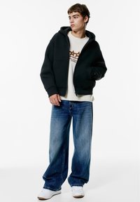 Jonge man die een zwarte capuchonjas draagt over een witte sweater met opdruk, wijde blauwe spijkerbroek en witte sneakers, staand tegen een effen witte achtergrond.