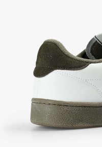 Zapatilla blanca con suela texturizada gris y cuello de talón de ante verde oliva oscuro, mostrando un detallado cosido y un forro interior acolchado.