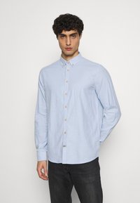 Camicia a maniche lunghe di colore azzurro chiaro in tessuto morbido, con colletto button-down e bottoni in legno, abbinata a jeans scuri.