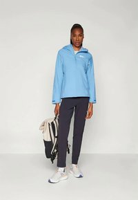Blue zip-up outdoor jacket, lightweight fabric, worn with black fitted pants and white sneakers. A beige and navy backpack rests nearby.