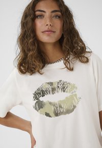 Krémové tričko s velkým stylizovaným potiskem rtů v zelené a černé barvě. Krátké rukávy, kulatý výstřih, hladký bavlněný povrch.