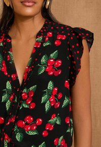 Schwarze Bluse mit rotem Kirschmuster und grünen Blättern; verfügt über rote Pünktchen-Details an den Ärmeln und ein V-Ausschnitt-Design.