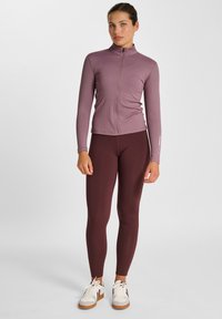 Langärmlige, zip-up Jacke aus leichtem lila geripptem Stoff, kombiniert mit bordeauxfarbenen Leggings. Das Outfit hat ein figurnahes Design und kontrastierende Sneakers.