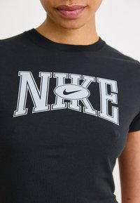 Črna bombažna majica s poudarjenim belim logotipom "NIKE" z detajlom v obliki kljuna, prilegajoč oblikovanjem, okroglim vratnim izrezom in kratkimi rokavi.
