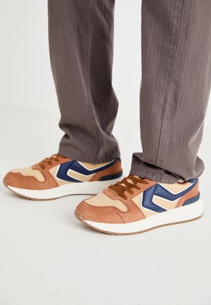 Brune og beige sneakers med marineblå accenter båret med brune bukser med nålestriber på et hvidt gulv.