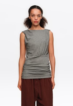 Mujer con cabello rizado que lleva una blusa gris sin mangas con fruncidos y pantalones marrones sueltos, de pie sobre un fondo blanco liso.