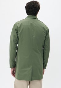 Abrigo ligero y verde con una abertura en la espalda, corte recto y mangas largas. Presenta un cuello clásico y una textura suave.