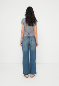 Γκρι καρό κοντό τοπ με λεπτομέρεια δέσιμου στο πίσω μέρος, συνδυασμένο με μπλε τζιν παντελόνι με φαρδιά πόδια και παπούτσια ανοιχτού χρώματος. Λευκό φόντο.