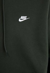 Tamsiai žalias džemperis su gobtuvu, pagamintas iš minkštos medžiagos, su baltu išsiuvinėtu Nike logotipu ant krūtinės ir raišteliu gobtuve.