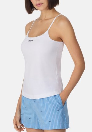 Frau trägt ein weißes Spaghettiträger-Top mit "DKNY"-Logo und hellblaue gestreifte Shorts mit kleinen "DKNY"-Logos, Hände in den Taschen.