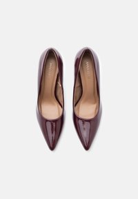 Par de sapatos de salto alto, de bico fino, em burgundy escuro brilhante, vistos de cima, com palmilhas de tom claro sobre um fundo branco.