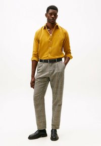 Uomo in piedi con le mani in tasca, che indossa una camicia gialla senape, pantaloni a quadri, cintura nera e scarpe nere su uno sfondo bianco.