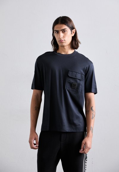 Ung mand med skulderlangt hår iført en sort t-shirt med en brystlomme og sorte bukser med hvid tekst på siden.