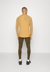 Hnědý top s dlouhým rukávem a kulatým výstřihem, doplněný olivovými legínami. Legíny mají zipy na kotnících.