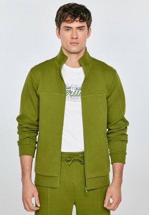 Młody mężczyzna w zielonym, zapinanym na zamek kurtce i spodniach, na białej koszulce z grafiką, stoi na jednolitym jasnym tle.