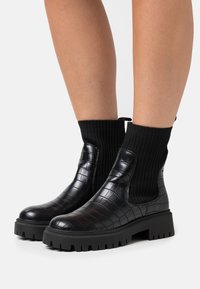 Botas negras de tobillo con textura de cocodrilo, caña elástica negra y suela gruesa. Presentan una parte superior tipo calcetín con canalé para un ajuste ceñido.