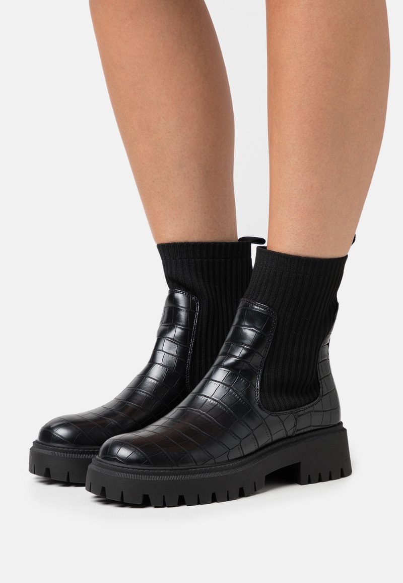 Botas negras de tobillo con textura de cocodrilo, caña elástica negra y suela gruesa. Presentan una parte superior tipo calcetín con canalé para un ajuste ceñido.
