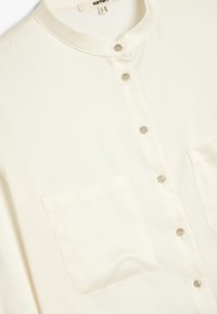 Blusa color crema realizzata in tessuto liscio, con colletto rotondo, chiusura con sei bottoni e due tasche frontali.