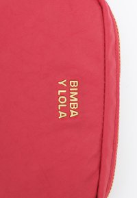 Czerwona tkaninowa sakwa z złotym tłoczonym logo "BIMBA Y LOLA", teksturowana powierzchnia i zamek błyskawiczny wzdłuż górnej krawędzi.