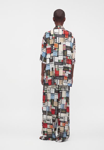 Personne portant une tenue ample assortie composée de deux pièces, recouverte d'imprimés graphiques rectangulaires colorés avec texte, debout, dos à l'appareil, sur un fond uni.