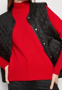 Gilet matelassé noir avec des boutons-pression sur un pull à col roulé rouge à côtes. Le tissu noir présente un motif en losange ; côtes au niveau des poignets et de l'ourlet.