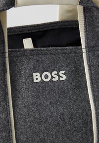 Siva torba za nošenje s bež trakama, koja na prednjoj strani ističe bijeli logo "BOSS". Minimalistički dizajn s glatkom teksturom.