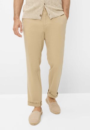 Mann trägt beige hochgekrempelte Chinos, passende Slipper und eine beige Strickjacke mit Knöpfen, eine Hand in der Tasche.