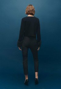 Suéter negro corto con puños forrados de piel, combinado con pantalones negros ajustados y tacones, frente a un fondo azul sólido.