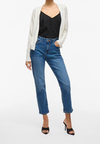 Cardigan bianco lavorato a maglia, top nero con spalline sottili, jeans blu a vita alta con taglio dritto e scarpe nere con tacco alto.