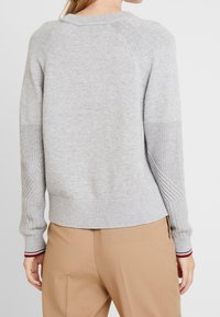 Woman wearing a light grey knit sweater with ribbed cuffs showing red, white, and black stripes, paired with beige pants, viewed from the back.