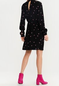 Robe noire à manches longues avec un petit motif floral rose. Détail en cœur au dos. Associée à des bottes de cheville rose vif et à des talons carrés.