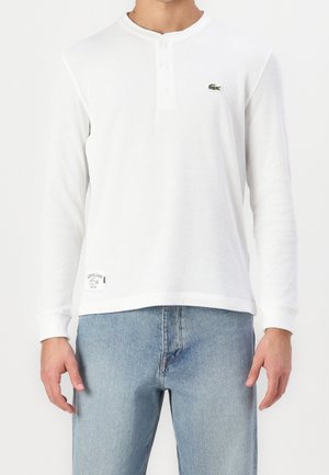 Hombre con camiseta henley blanca de manga larga con tres botones, pequeño logo de cocodrilo en el pecho y vaqueros azul claro.