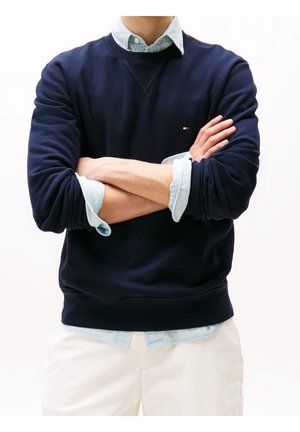 Person trägt marineblaue Sweatshirt über hellblauem Hemd mit Knöpfen, Arme verschränkt und weiße Hose, vor einfarbigem Hintergrund.