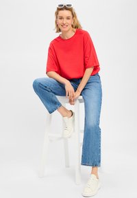 Rode oversized t-shirt met korte mouwen, gecombineerd met lichtblauwe rechte jeans en witte sneakers, zittend op een witte kruk.