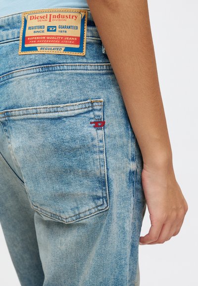 Des jeans en denim bleu clair présentent une étiquette de marque "Diesel Industry" visible, des coutures rouges sur la poche et un design fuselé avec une texture décolorée.
