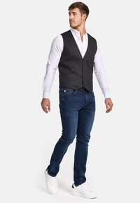 Grau strukturiertes Weste über einem weißen Hemd, kombiniert mit dunkelblauen Jeans und weißen Sneakers. Die Weste hat eine taillierte Form und Knöpfe.