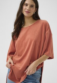 Terasz színű, oversize pamut póló, puha, texturált anyagból készült, kerek nyakkivágással és rövid ujjal, farmernadrággal viselve.