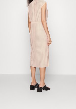 Femme portant une robe midi plissée rose clair sans manches et des sandales noires à talons blocs et bouts ouverts, debout de dos.