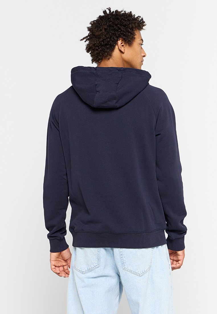 Felpa blu navy con una texture morbida, caratterizzata da un cappuccio con cordini e polsini a costine. Il design include una vestibilità rilassata e dettagli minimi.