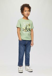 Groen T-shirt met korte mouwen met een afbeelding van een gele graafmachine, gecombineerd met blauwe spijkerbroeken en witte sneakers met grijze accenten.