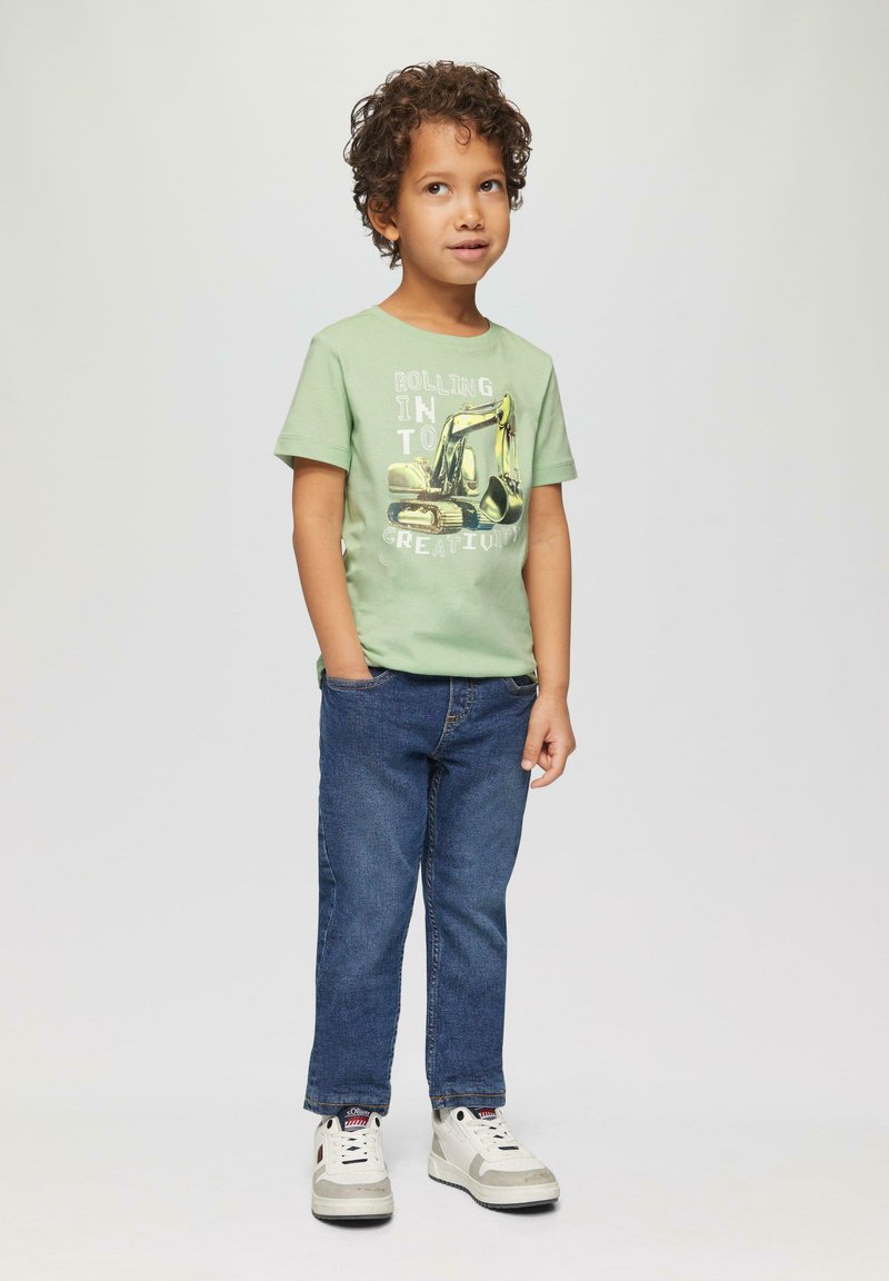 Groen T-shirt met korte mouwen met een afbeelding van een gele graafmachine, gecombineerd met blauwe spijkerbroeken en witte sneakers met grijze accenten.