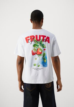 Mann trägt weißes T-Shirt mit großem roten Schriftzug "FRUTA Fresca" und Grafik von Erdbeeren und grünen Blättern auf dem Rücken.