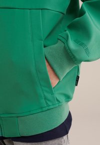 Felpa verde con una superficie liscia, dotata di una tasca frontale, polsini a costine e un strato inferiore blu navy in contrasto. Dettaglio di branding sottile visibile.