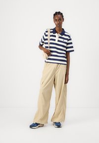 Polo a righe blu navy e bianche con colletto, abbinato a pantaloni cargo beige larghi e scarpe da ginnastica blu. Include una piccola borsa a tracolla.
