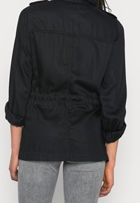 Sort jakke med indsnævret talje, lange ærmer op-rullet og rygdetalje. Fremstillet af letvægtsstof med en glat tekstur.