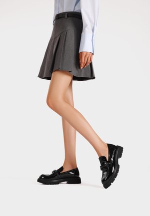 Scarpe in vernice nera con suola chunky, dotate di dettaglio con cinturino e fibbia, indossate con una gonna grigia e una blusa blu.