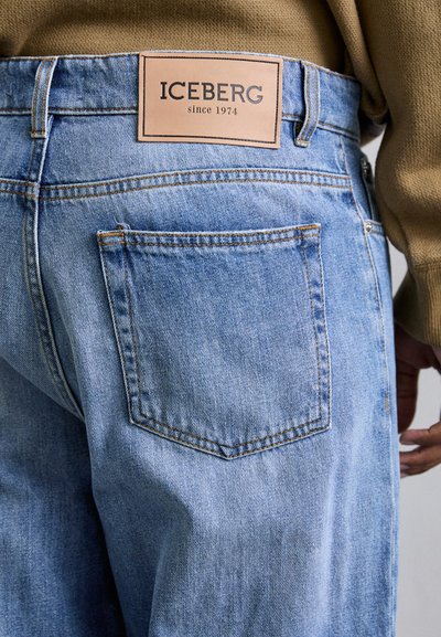 Lys blå denim jeans med et lædermærke, der viser "ICEBERG siden 1974", med almindelige baglommer og orange syninger.