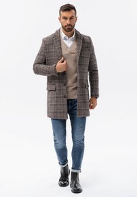 Ruskea ruudullinen päällystakki, jossa on notch-lapelit ja nappikiinnitys, käytetty vaaleanbeigen neuleen päällä, yhdistetty sinisiin farkkuihin ja mustiin nilkkasaappaisiin.