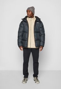 Chaqueta de plumón negra con capucha sobre una chaqueta de forro polar beige, combinada con pantalones negros y un gorro gris. Los zapatos son de un gris claro con detalles oscuros.