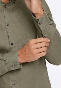Camisa de lino verde con botones en la parte delantera y un bolsillo en el pecho. La tela tiene un acabado texturizado, y la manga está ligeramente arremangada.